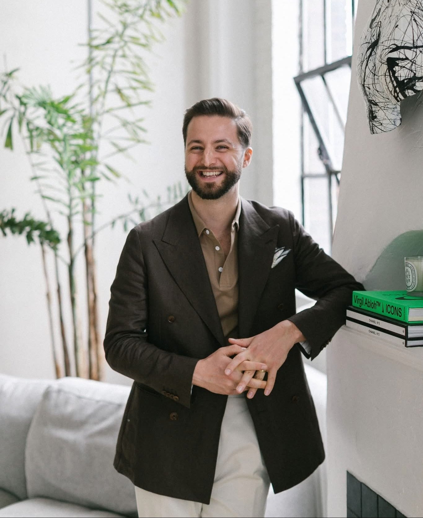 A smiling man with a beard wearing a dark brown double-breasted blazer over a tan button-down shirt and cream trousers, standing in a bright, modern room with a sofa, potted plant, and window.